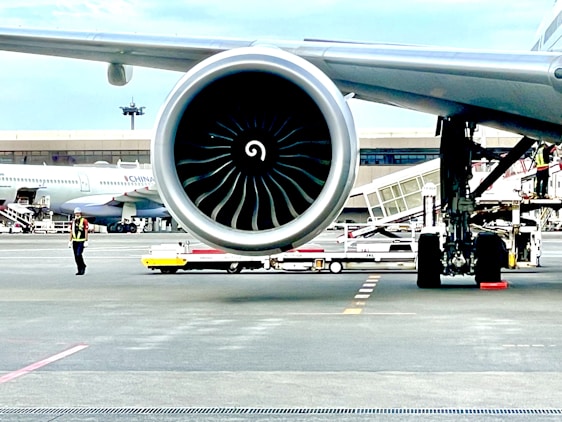 A close-up view of a large aircraft jet engine on a runway with a terminal in the background. An airport worker in a reflective vest and helmet is visible nearby, along with other airport equipment and vehicles. Another plane is parked in the distance.