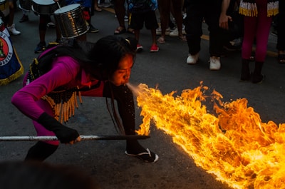 A performer is displaying a fire-breathing act in the middle of a street surrounded by onlookers. The individual is wearing a vibrant magenta outfit with decorative elements around the waist. Flames are dramatically extending forward, illuminating the scene against the dim asphalt background. Musicians with drums are part of the crowd, contributing to a festive atmosphere.