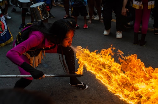 A performer is displaying a fire-breathing act in the middle of a street surrounded by onlookers. The individual is wearing a vibrant magenta outfit with decorative elements around the waist. Flames are dramatically extending forward, illuminating the scene against the dim asphalt background. Musicians with drums are part of the crowd, contributing to a festive atmosphere.