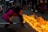 A performer is displaying a fire-breathing act in the middle of a street surrounded by onlookers. The individual is wearing a vibrant magenta outfit with decorative elements around the waist. Flames are dramatically extending forward, illuminating the scene against the dim asphalt background. Musicians with drums are part of the crowd, contributing to a festive atmosphere.