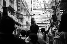 A busy urban street scene features a group of people gathered, many holding up smartphones to take photos. The scene is characterized by a tangle of overhead wires and a backdrop of tall buildings with signage in another language. The composition includes a cyclist wearing a helmet, which stands out among the crowd.