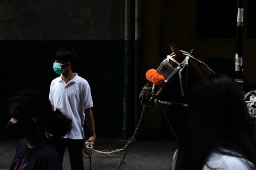 A person wearing a surgical mask and a white polo shirt is holding the reins of a horse adorned with a vibrant orange pom-pom on its bridle. The setting appears to be dimly lit with a dark background, creating a contrast with the bright outfit and the horse's decorations. Several other people are partially visible in the foreground.