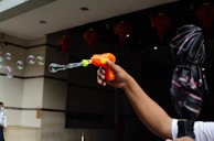 A person holds an orange toy bubble gun, releasing delicate bubbles into the air. In the background, red lanterns are partially visible, and to the side, someone stands near a building's entrance.