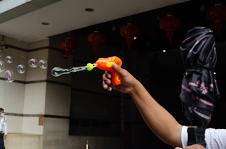 A person holds an orange toy bubble gun, releasing delicate bubbles into the air. In the background, red lanterns are partially visible, and to the side, someone stands near a building's entrance.