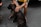A person is seated on a chair, wearing a yellow shirt and casual shorts, holding a dark-colored dog on a leash. The scene is set on a paved surface, and the person is wearing black sliders.