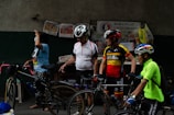 Several adults are standing beside bicycles, wearing cycling helmets and gear. There are various newspapers and bags pinned to a wall labeled 'Barangay Livelihood Program.' The atmosphere appears casual and relaxed.