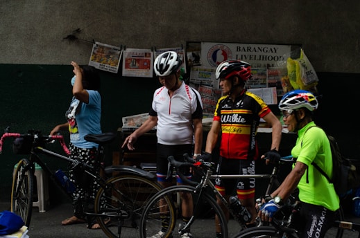Several adults are standing beside bicycles, wearing cycling helmets and gear. There are various newspapers and bags pinned to a wall labeled 'Barangay Livelihood Program.' The atmosphere appears casual and relaxed.