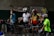 Several adults are standing beside bicycles, wearing cycling helmets and gear. There are various newspapers and bags pinned to a wall labeled 'Barangay Livelihood Program.' The atmosphere appears casual and relaxed.