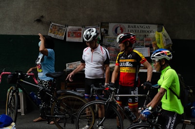 Several adults are standing beside bicycles, wearing cycling helmets and gear. There are various newspapers and bags pinned to a wall labeled 'Barangay Livelihood Program.' The atmosphere appears casual and relaxed.