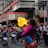 A street performer breathes fire surrounded by a group of people, including drummers and onlookers. The setting appears to be a lively street festival with red lanterns hanging above and colorful banners on display.