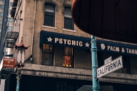 A street scene with a psychic shop located on a corner. The shop features a navy blue awning with the words 'Psychic Palm & Tarot' displayed. There are two windows, one of which has a sign in neon lights advertising services related to predicting the past, present, and future. A traditional street lamp with a decorative topper adds an element of cultural design. The image also shows a street sign for California Street and nearby buildings, including emergency stairs.