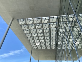 The photo captures a modern architectural structure featuring large overhanging concrete slabs supported by tall, slender concrete columns. The ceiling has a grid-like pattern with square openings allowing sunlight to pass through, creating a geometric shadow on the surface. Adjacent to this structure, there is a building facade with large glass windows that reflect the sky and surrounding environment.