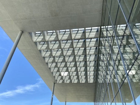 The photo captures a modern architectural structure featuring large overhanging concrete slabs supported by tall, slender concrete columns. The ceiling has a grid-like pattern with square openings allowing sunlight to pass through, creating a geometric shadow on the surface. Adjacent to this structure, there is a building facade with large glass windows that reflect the sky and surrounding environment.
