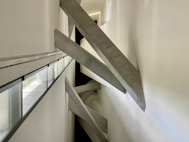 Concrete beams intersect diagonally within a narrow, modern architectural space. The design creates a dramatic visual effect with sharp angles and strong geometric lines. Natural light filters in through long, narrow windows on the left side, casting subtle shadows on the white walls.