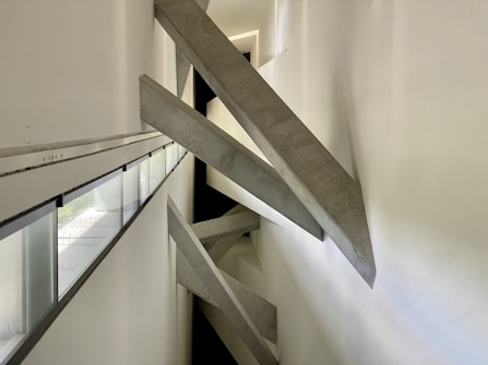 Concrete beams intersect diagonally within a narrow, modern architectural space. The design creates a dramatic visual effect with sharp angles and strong geometric lines. Natural light filters in through long, narrow windows on the left side, casting subtle shadows on the white walls.