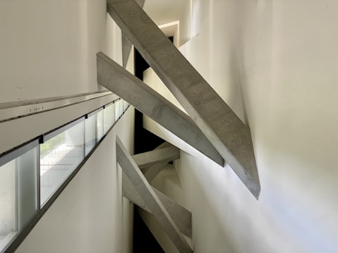 Concrete beams intersect diagonally within a narrow, modern architectural space. The design creates a dramatic visual effect with sharp angles and strong geometric lines. Natural light filters in through long, narrow windows on the left side, casting subtle shadows on the white walls.