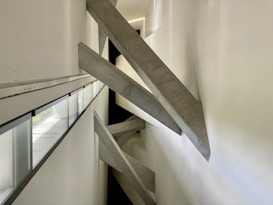 Concrete beams intersect diagonally within a narrow, modern architectural space. The design creates a dramatic visual effect with sharp angles and strong geometric lines. Natural light filters in through long, narrow windows on the left side, casting subtle shadows on the white walls.