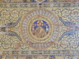 An intricate mosaic artwork featuring a central figure, possibly a religious icon, surrounded by elaborate patterns and text. The design includes floral motifs, geometric shapes, and repeating symbols, with a surrounding circular frame.