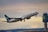 A commercial airplane is taking off, with its nose pointed upward. The aircraft is painted in the colors and logo of WestJet. In the background, there is an airport control tower and a sky filled with clouds displaying various hues, suggesting either dawn or dusk.