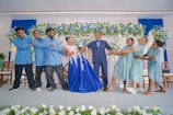 A group of people is dressed in coordinated attire, participating in a playful moment at a celebration, likely a wedding. The central figure, a woman in a vibrant blue gown, is being pulled from both sides by different groups of people; men are dressed in matching blue shirts with patterns and women in light blue dresses. The backdrop features an elaborate arrangement of flowers and blue and white drapery.