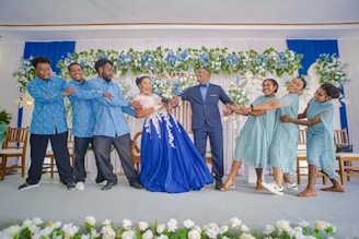 A group of people is dressed in coordinated attire, participating in a playful moment at a celebration, likely a wedding. The central figure, a woman in a vibrant blue gown, is being pulled from both sides by different groups of people; men are dressed in matching blue shirts with patterns and women in light blue dresses. The backdrop features an elaborate arrangement of flowers and blue and white drapery.
