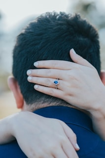 A woman's hand, adorned with a sapphire ring, gently rests on the back of a man's head. Her nails are neatly manicured in a light shade. The man is wearing a dark blue shirt, and there is a sense of intimacy and connection between the two people.