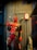 A large, detailed statue of a superhero character wearing a red and black costume with two swords strapped to the back. The figure is standing in a confident pose with one finger raised, situated in a store environment with merchandise visible on the shelves.