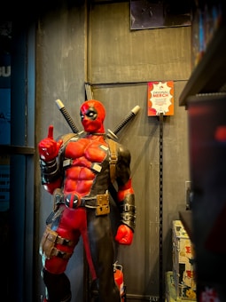 A large, detailed statue of a superhero character wearing a red and black costume with two swords strapped to the back. The figure is standing in a confident pose with one finger raised, situated in a store environment with merchandise visible on the shelves.