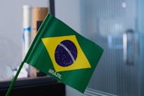 A small Brazilian flag with the green field, yellow rhombus, and blue globe with stars, featuring the motto 'Ordem e Progresso' in a white band across the globe. The flag is mounted on a green pole and is placed in an indoor setting.
