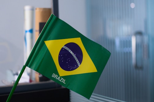 A small Brazilian flag with the green field, yellow rhombus, and blue globe with stars, featuring the motto 'Ordem e Progresso' in a white band across the globe. The flag is mounted on a green pole and is placed in an indoor setting.