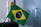A small Brazilian flag with the green field, yellow rhombus, and blue globe with stars, featuring the motto 'Ordem e Progresso' in a white band across the globe. The flag is mounted on a green pole and is placed in an indoor setting.