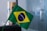 A small Brazilian flag with the green field, yellow rhombus, and blue globe with stars, featuring the motto 'Ordem e Progresso' in a white band across the globe. The flag is mounted on a green pole and is placed in an indoor setting.