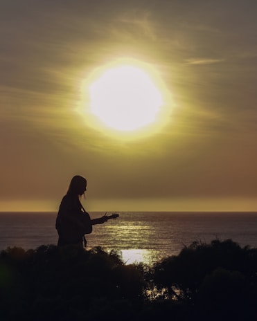 A serene sunset with a silhouette of a person playing guitar by the water.