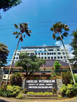 A university building is visible behind a large sign that reads 'Fakultas Peternakan (Faculty of Animal Science) Universitas Brawijaya.' The building is several stories tall, with numerous windows. There are two tall palm trees and other greenery, including bushes and plants, in the foreground. The sky is clear and bright blue with some scattered clouds.