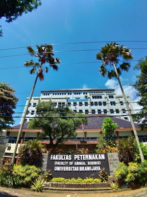 A university building is visible behind a large sign that reads 'Fakultas Peternakan (Faculty of Animal Science) Universitas Brawijaya.' The building is several stories tall, with numerous windows. There are two tall palm trees and other greenery, including bushes and plants, in the foreground. The sky is clear and bright blue with some scattered clouds.
