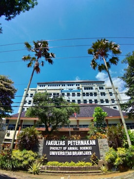 A university building is visible behind a large sign that reads 'Fakultas Peternakan (Faculty of Animal Science) Universitas Brawijaya.' The building is several stories tall, with numerous windows. There are two tall palm trees and other greenery, including bushes and plants, in the foreground. The sky is clear and bright blue with some scattered clouds.
