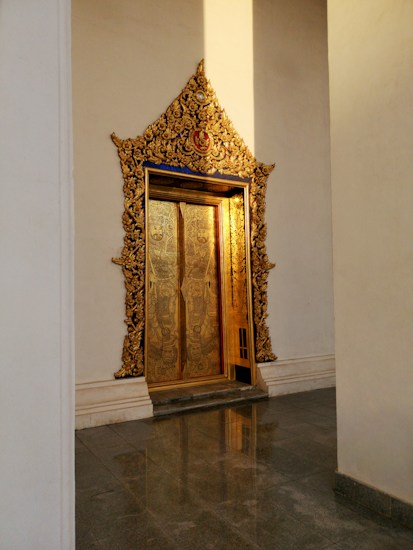 A beautifully ornate doorway featuring intricate gold detailing and patterns. The door appears to be made from metal with elaborate engravings and is set within a tall, arched frame that is also richly decorated with golden embellishments. The sunlight casts a warm glow on the door, enhancing its luxurious appearance. The surrounding walls are plain, creating a striking contrast with the door's opulence.