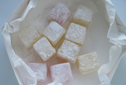 A collection of small, square-shaped candies coated in powdered sugar, arranged on a piece of crumpled parchment paper. The candies appear in pale pastel shades, including pink and white.