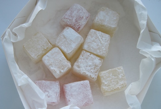A collection of small, square-shaped candies coated in powdered sugar, arranged on a piece of crumpled parchment paper. The candies appear in pale pastel shades, including pink and white.
