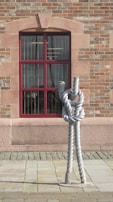 A large, intricately knotted rope sculpture stands in front of a wall made of red and tan bricks. The wall features a window with a red frame, through which indoor lights and faint images are visible. The sculpture is positioned on a cobblestone pavement.