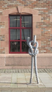 A large, intricately knotted rope sculpture stands in front of a wall made of red and tan bricks. The wall features a window with a red frame, through which indoor lights and faint images are visible. The sculpture is positioned on a cobblestone pavement.
