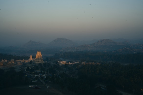 Aerial view of an ancient temple complex at dawn, with a large temple tower in the center surrounded by smaller structures. The landscape includes distant rolling hills under a hazy sky, and dense greenery in the foreground.