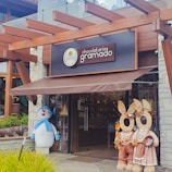 A storefront of a chocolate shop named 'Chocolataria Gramado' with a pergola-style wooden structure above. Two large plush figures resembling rabbits and a snowman stand near the entrance, adding a whimsical touch.