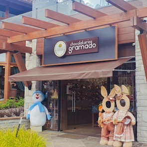 A storefront of a chocolate shop named 'Chocolataria Gramado' with a pergola-style wooden structure above. Two large plush figures resembling rabbits and a snowman stand near the entrance, adding a whimsical touch.