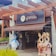 A storefront of a chocolate shop named 'Chocolataria Gramado' with a pergola-style wooden structure above. Two large plush figures resembling rabbits and a snowman stand near the entrance, adding a whimsical touch.