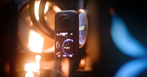 A highly detailed close-up of a technological device with a screen displaying the text 'NZXT.' The background has a mixture of warm and cool lighting, with some blurred elements, including fan-like structures.