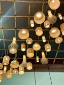A ceiling display features multiple hanging bamboo lamps with unique woven designs, enhanced by warm, glowing light bulbs. The lamps vary in shape and size, adding an intricate and artistic flair to the setting. The background consists of a wooden grid ceiling that complements the rustic look.