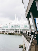 A waterfront scene featuring a modern building with large glass windows and white peaked roofs. People walk along the railing of a promenade that overlooks the water. The word 'CANADA' is marked prominently on the building, and the structure appears to be part of a large complex, likely a convention center.