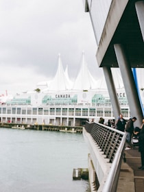 A waterfront scene featuring a modern building with large glass windows and white peaked roofs. People walk along the railing of a promenade that overlooks the water. The word 'CANADA' is marked prominently on the building, and the structure appears to be part of a large complex, likely a convention center.