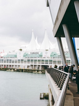 A waterfront scene featuring a modern building with large glass windows and white peaked roofs. People walk along the railing of a promenade that overlooks the water. The word 'CANADA' is marked prominently on the building, and the structure appears to be part of a large complex, likely a convention center.
